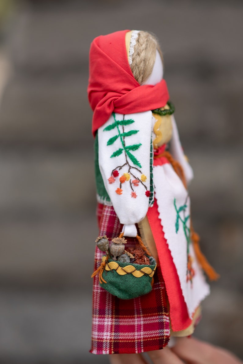Puede incluir: Una mu&ntilde;eca tradicional folcl&oacute;rica con un pa&ntilde;uelo rojo, una blusa blanca bordada y una falda de cuadros rojos y negros. La mu&ntilde;eca lleva una cesta verde con flores secas.