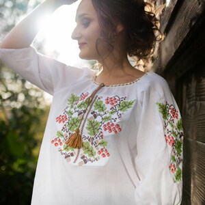 May include: A white embroidered blouse with a floral design featuring red berries and green leaves. The blouse has a tassel detail on the neckline.