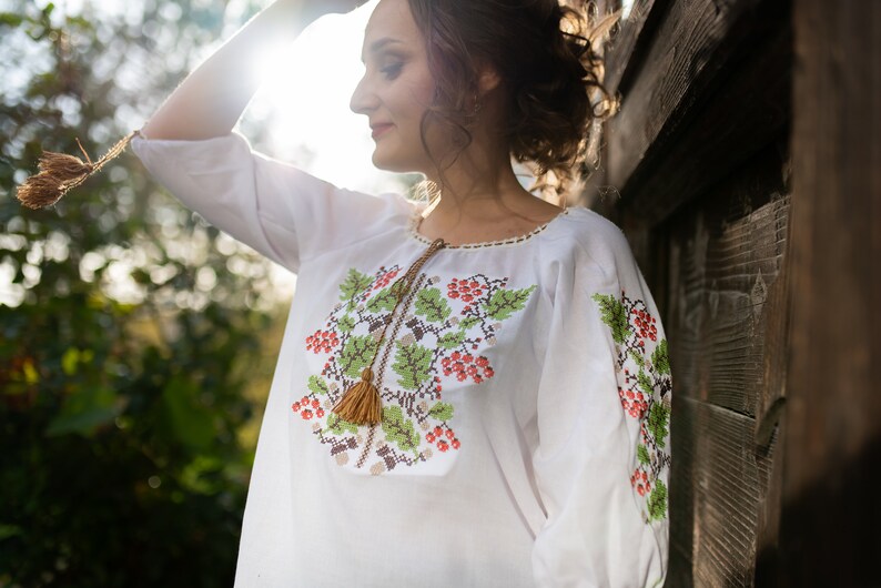 May include: A white embroidered blouse with a floral design featuring red berries and green leaves. The blouse has a tassel detail on the neckline.