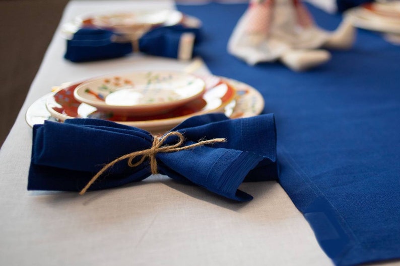 May include: A table setting featuring a navy blue linen napkin tied with twine, placed near a decorative plate with a floral design. A blue tablecloth and white tablecloth are also visible, creating a layered effect.