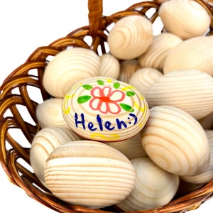 K&ouml;nnte beinhalten: Ein Weidenkorb gef&uuml;llt mit Holzeiern. Ein Ei ist mit einem rot-gr&uuml;nen Blumenmuster und dem Namen "Helen" in Blau bemalt. Der Text "The painting is made as an example" ist sichtbar.