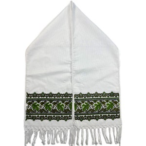 May include: White linen table runner with a green and dark green leaf pattern border and fringe. The runner is folded in half lengthwise, with a pointed top. The leaf pattern is repeated along the bottom edge.