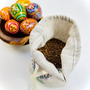 K&ouml;nnte beinhalten: Eine Holzschale enth&auml;lt mehrere bunt dekorierte Ostereier. Die Eier weisen aufwendige Muster in Orange, Gelb, Gr&uuml;n und Blau auf. Daneben liegt ein kleiner, offener Leinensack, gef&uuml;llt mit braunen K&ouml;rnern, mit einem wei&szlig;en Band gebunden.