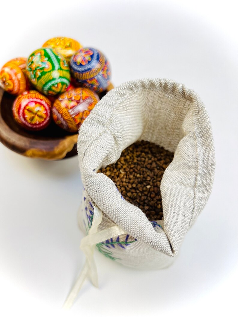 K&ouml;nnte beinhalten: Eine Holzschale enth&auml;lt mehrere bunt dekorierte Ostereier. Die Eier weisen aufwendige Muster in Orange, Gelb, Gr&uuml;n und Blau auf. Daneben liegt ein kleiner, offener Leinensack, gef&uuml;llt mit braunen K&ouml;rnern, mit einem wei&szlig;en Band gebunden.