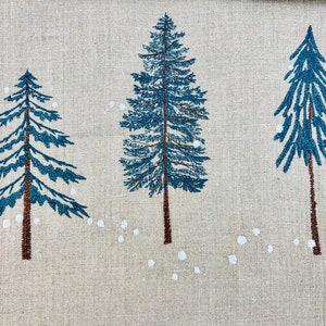 Op de afbeelding: Een beige linnen stof met drie blauwe geborduurde dennenbomen en witte geborduurde sneeuwvlokken.