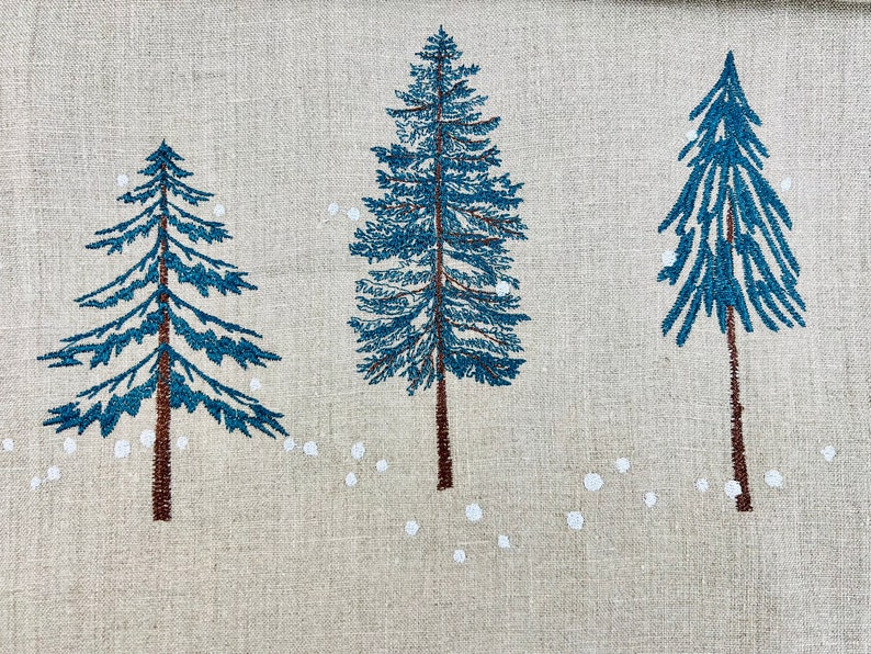 Op de afbeelding: Een beige linnen stof met drie blauwe geborduurde dennenbomen en witte geborduurde sneeuwvlokken.