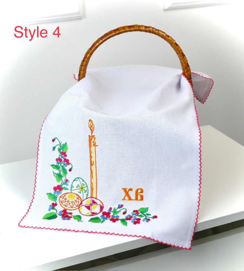 K&ouml;nnte beinhalten: Wei&szlig;er Leinen-Osterk&ouml;rbchen-Einsatz mit gestickten roten, gelben und gr&uuml;nen Blumen, einer Kerze und drei verzierten Eiern. Der Einsatz hat einen rosa gewellten Rand und der Text "XB" ist in Orange gestickt.