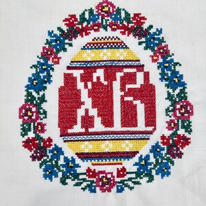 Pu&ograve; includere: Un tessuto bianco con un ricamo colorato a forma di uovo di Pasqua circondato da una ghirlanda di fiori. L'uovo &egrave; rosso, giallo e blu con un'iscrizione bianca all'interno.