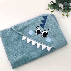 May include: A light blue hooded towel with a white and blue embroidered dinosaur face. The dinosaur has two large eyes, a wide mouth with white teeth, and a blue fin on top of its head.