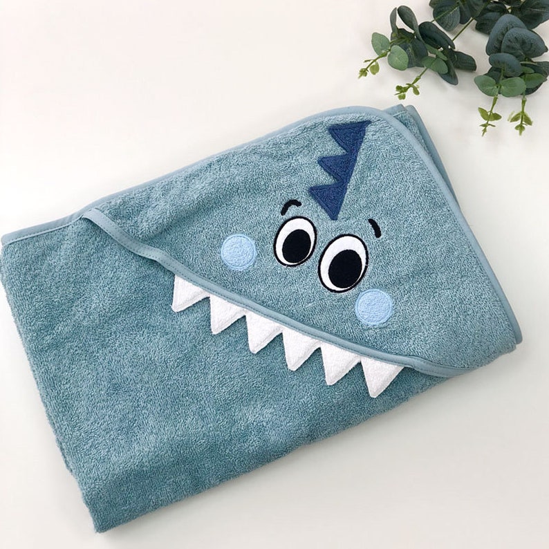 May include: A light blue hooded towel with a white and blue embroidered dinosaur face. The dinosaur has two large eyes, a wide mouth with white teeth, and a blue fin on top of its head.