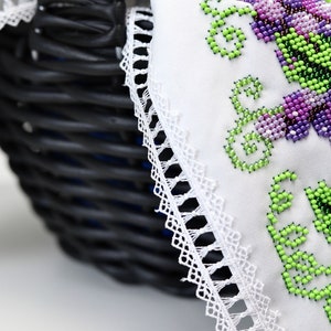 Puede incluir: Una cesta de mimbre negra con un forro de tela blanca con bordados florales de cuentas en tonos verdes y morados. El borde de la tela est&aacute; adornado con un delicado encaje blanco, creando una est&eacute;tica artesanal.