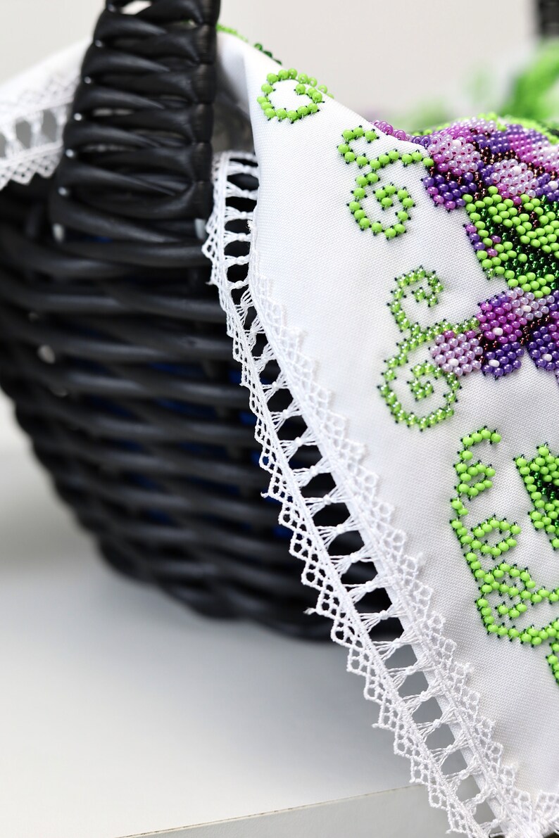 Puede incluir: Una cesta de mimbre negra con un forro de tela blanca con bordados florales de cuentas en tonos verdes y morados. El borde de la tela est&aacute; adornado con un delicado encaje blanco, creando una est&eacute;tica artesanal.