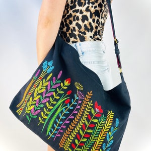 K&ouml;nnte beinhalten: Eine schwarze Canvas-Tragetasche mit einem farbenfrohen, gestickten Blumenmuster. Die Blumen sind in Rot-, Orange-, Gelb-, Gr&uuml;n-, Blau- und Violettt&ouml;nen gehalten.