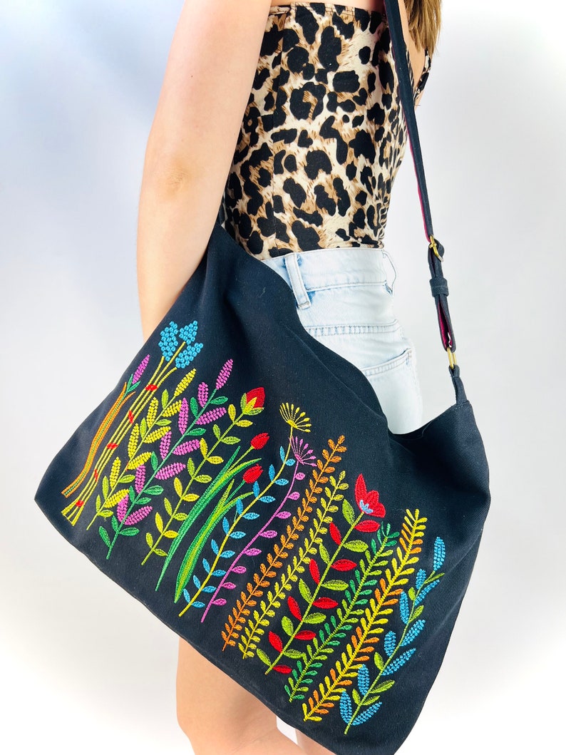 K&ouml;nnte beinhalten: Eine schwarze Canvas-Tragetasche mit einem farbenfrohen, gestickten Blumenmuster. Die Blumen sind in Rot-, Orange-, Gelb-, Gr&uuml;n-, Blau- und Violettt&ouml;nen gehalten.