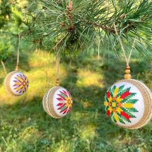 May include: Three handmade Christmas ornaments with a burlap base and colorful embroidered designs. The ornaments are hanging from a pine tree branch.