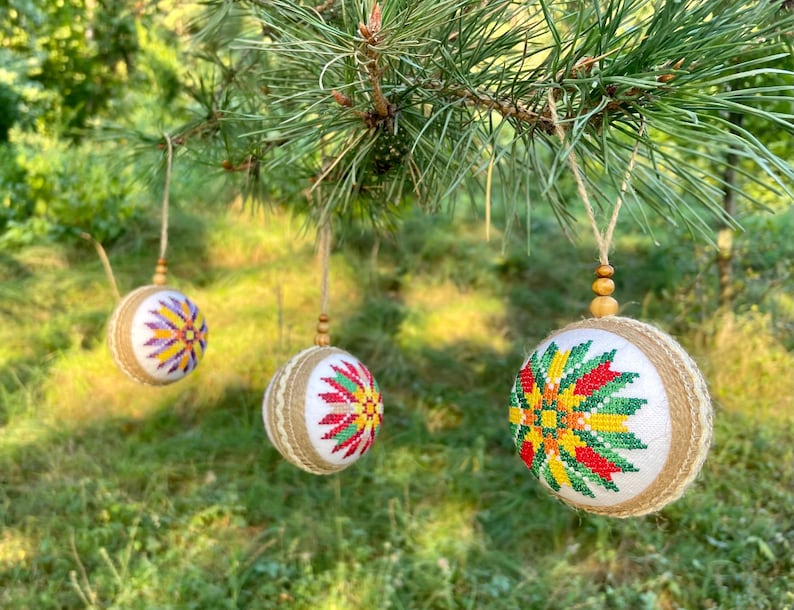 May include: Three handmade Christmas ornaments with a burlap base and colorful embroidered designs. The ornaments are hanging from a pine tree branch.