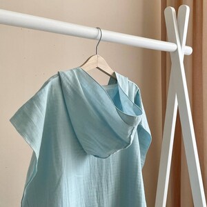 Puede incluir: Un poncho de lino azul claro con capucha colgado de una percha de madera blanca. El poncho tiene un corte holgado y un dise&ntilde;o sencillo.