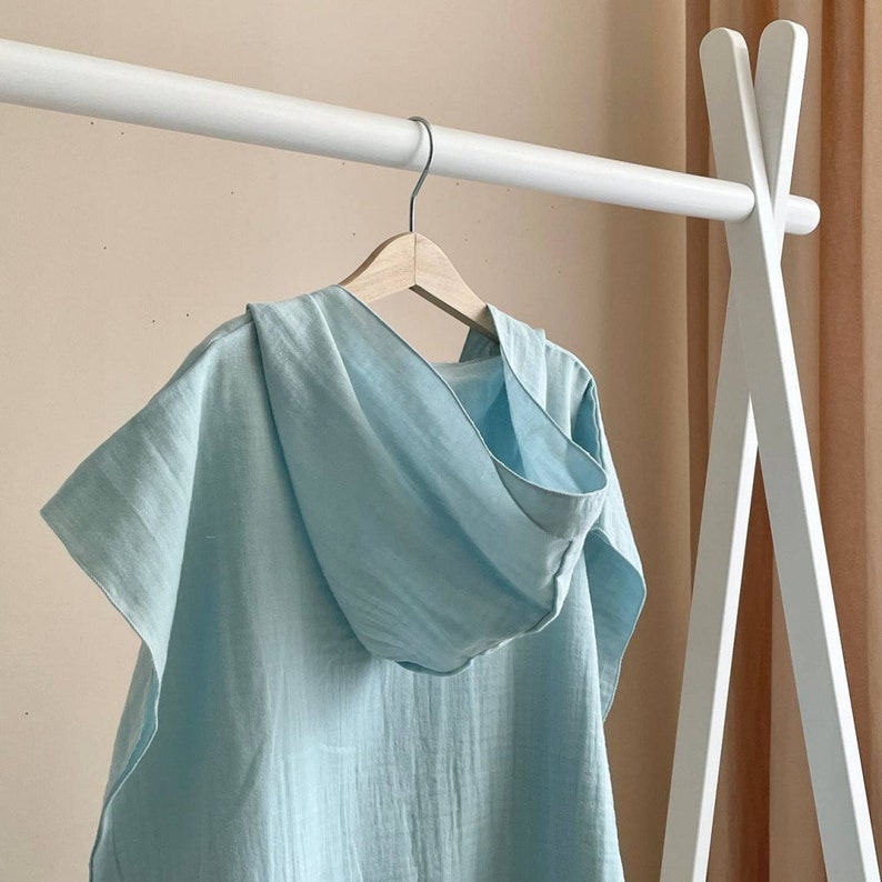 Puede incluir: Un poncho de lino azul claro con capucha colgado de una percha de madera blanca. El poncho tiene un corte holgado y un dise&ntilde;o sencillo.