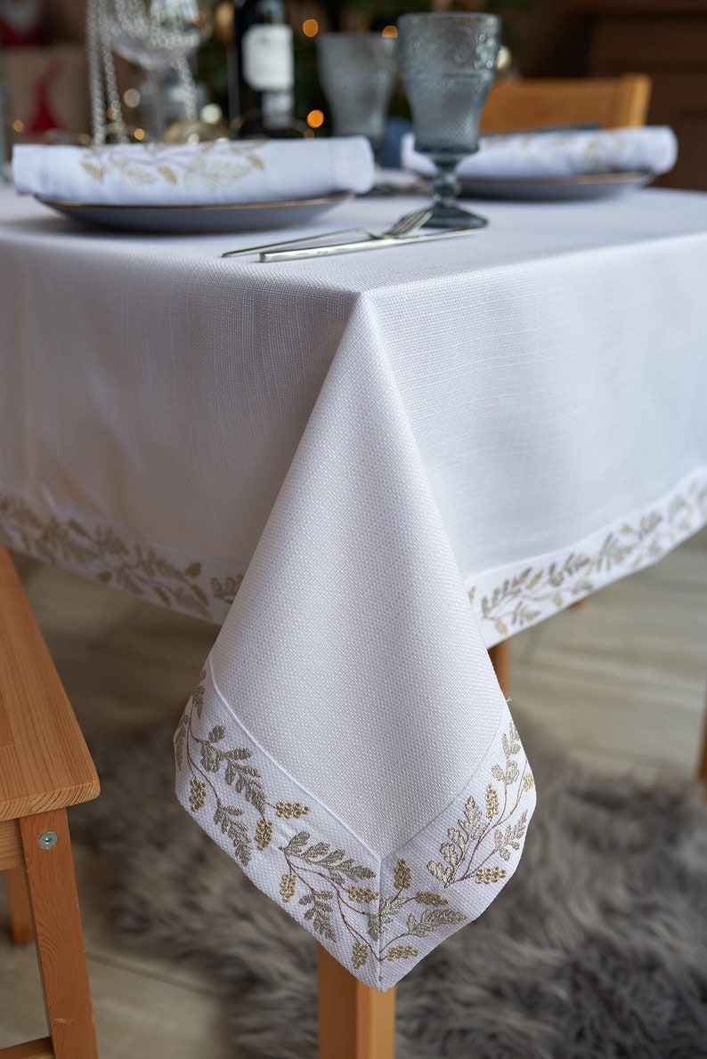 Puede incluir: Mantel de lino blanco con un borde bordado con hojas doradas. La mesa est&aacute; puesta con servilletas, platos y cubiertos a juego, creando un ambiente elegante.