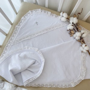 May include: White baby blanket with a delicate lace trim and a small embroidered cross in the center. The blanket is folded in half and laid on a white crib mattress.