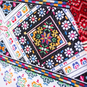 May include: Close-up of a white fabric with colorful embroidered patterns. The design features geometric shapes and floral motifs in red, blue, green, yellow, and black.