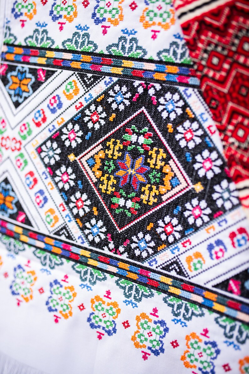 May include: Close-up of a white fabric with colorful embroidered patterns. The design features geometric shapes and floral motifs in red, blue, green, yellow, and black.