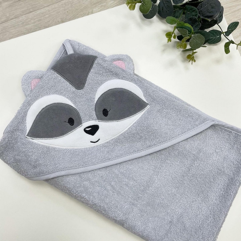 May include: A grey hooded baby towel with a raccoon face design. The towel features a grey hood with a dark grey patch, white and grey eyes, and pink inner ears. The towel is made of soft material.