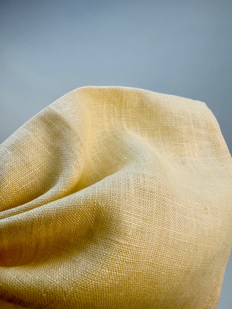 Puede incluir: Primer plano de una tela de lino beige con un tejido texturizado. La tela est&aacute; drapeada, creando pliegues y sombras suaves. El color es un tono c&aacute;lido y natural, lo que sugiere textiles y decoraci&oacute;n del hogar.