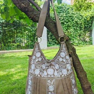 K&ouml;nnte beinhalten: Eine taupefarbene Umh&auml;ngetasche mit wei&szlig;er Blumenstickerei. Die Tasche hat ein plissiertes Design und lange Riemen, die an goldfarbenen Ringen befestigt sind. Die Tasche h&auml;ngt an einem Ast vor dem Hintergrund von gr&uuml;nem Gras und Bl&auml;ttern.