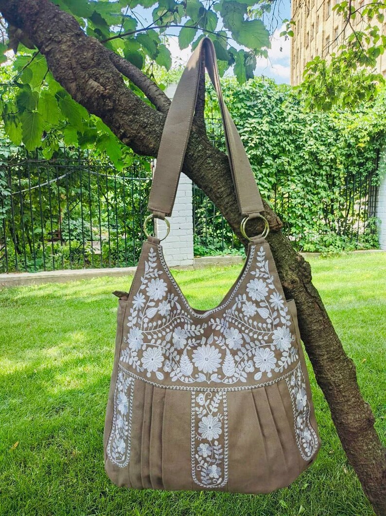 K&ouml;nnte beinhalten: Eine taupefarbene Umh&auml;ngetasche mit wei&szlig;er Blumenstickerei. Die Tasche hat ein plissiertes Design und lange Riemen, die an goldfarbenen Ringen befestigt sind. Die Tasche h&auml;ngt an einem Ast vor dem Hintergrund von gr&uuml;nem Gras und Bl&auml;ttern.