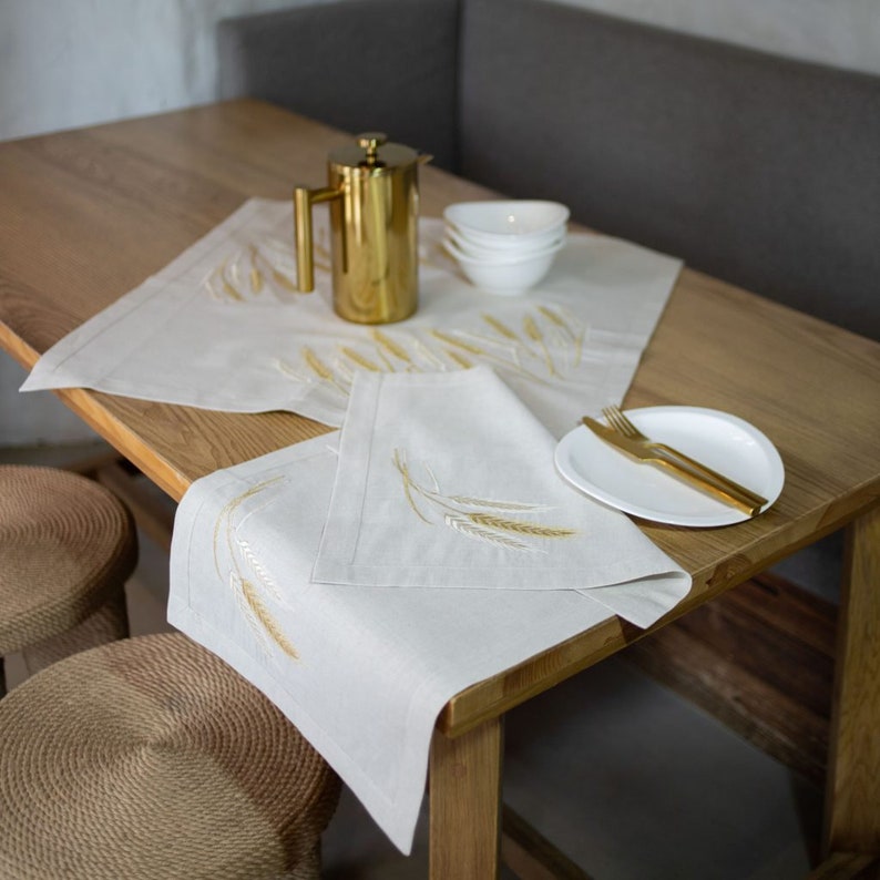 Peut inclure: Une table de salle &agrave; manger avec une cafeti&egrave;re dor&eacute;e, des bols blancs et une assiette blanche avec des couverts dor&eacute;s. Deux serviettes de table en lin blanc cass&eacute; et un chemin de table sont brod&eacute;s d'&eacute;pis de bl&eacute; dor&eacute;s.