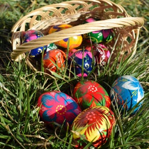 May include: A wicker basket overflowing with vibrantly painted Easter eggs rests in green grass. The eggs feature intricate floral designs in red, blue, yellow, and pink. The basket's handle arches over the colorful display.