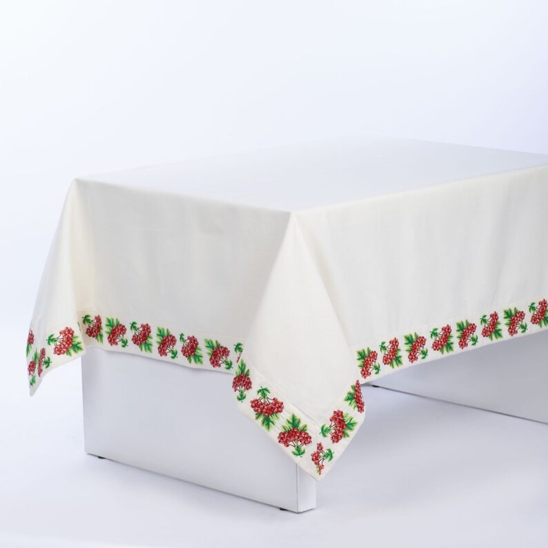 May include: Off-white tablecloth with a decorative border featuring embroidered red berries and green leaves. The tablecloth is draped over a white surface, showcasing the detailed floral design. A classic piece for dining.