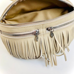 Puede incluir: Ri&ntilde;onera color crema con cierre de cremallera y dise&ntilde;o de flecos. La bolsa tiene dos compartimentos con cremallera y una cremallera plateada. Los tiradores de la cremallera tienen forma de hoja. El interior de la bolsa es de color beige claro.