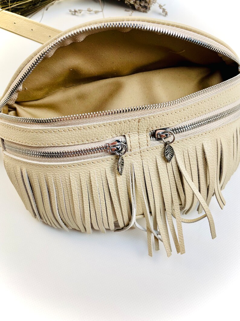 Puede incluir: Ri&ntilde;onera color crema con cierre de cremallera y dise&ntilde;o de flecos. La bolsa tiene dos compartimentos con cremallera y una cremallera plateada. Los tiradores de la cremallera tienen forma de hoja. El interior de la bolsa es de color beige claro.