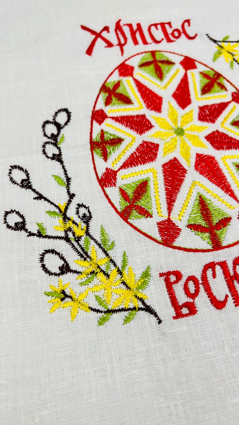 May include: Embroidered white fabric with a large, decorated Easter egg in red, yellow, and green. The text "Christ is Risen" is embroidered in red. Floral and branch embroidery in yellow and green.