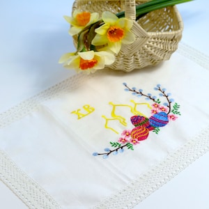 May include: A wicker basket with a handle contains daffodils with yellow and orange flowers. A white embroidered cloth with a lace border features colourful Easter egg designs and the letters "XB".