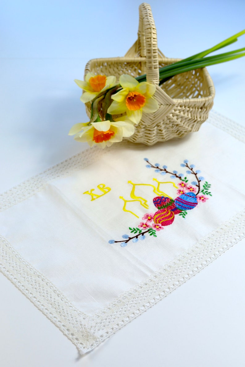 May include: A wicker basket with a handle contains daffodils with yellow and orange flowers. A white embroidered cloth with a lace border features colourful Easter egg designs and the letters "XB".