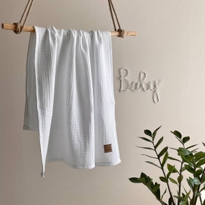 K&ouml;nnte beinhalten: Eine wei&szlig;e Babydecke aus Musselin h&auml;ngt an einer Holzstange, die an Seilen befestigt ist. Ein kleines braunes Lederetikett ist an der Decke befestigt. Das Wort "Baby" steht in Drahtschrift an der Wand.