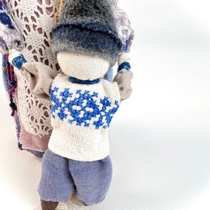 May include: A handmade rag doll with a blue and white embroidered shirt, blue pants, and a gray hat. The doll is holding the hand of another doll, which is partially visible.
