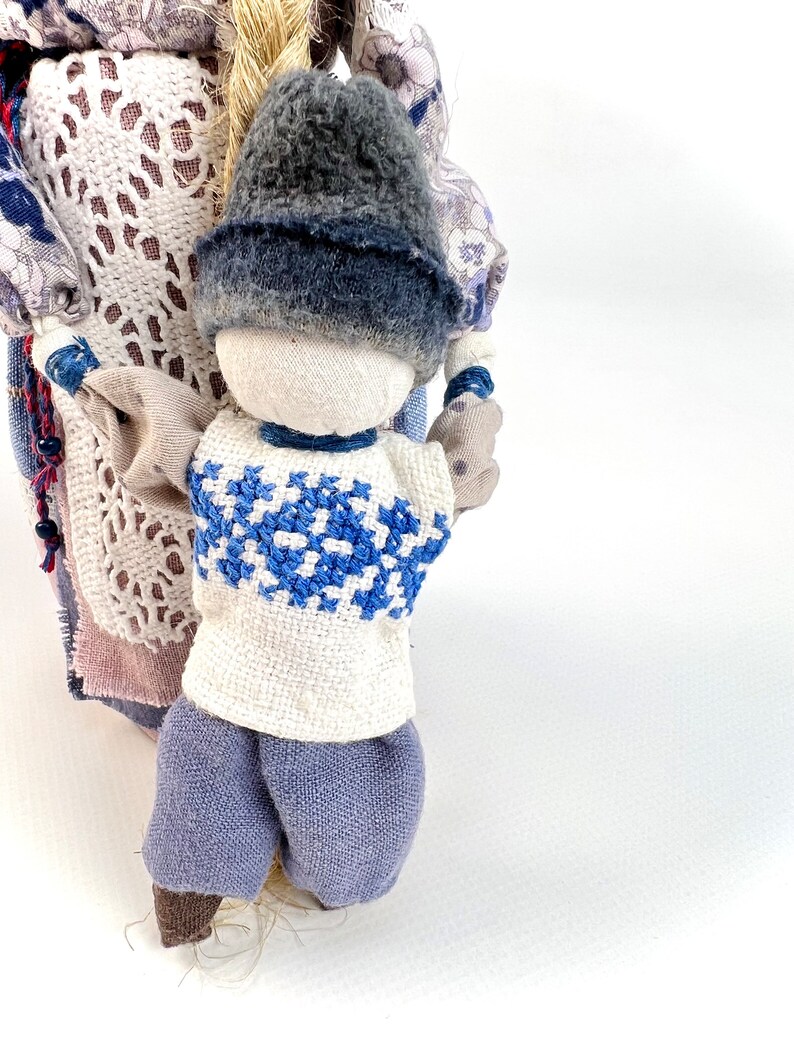 May include: A handmade rag doll with a blue and white embroidered shirt, blue pants, and a gray hat. The doll is holding the hand of another doll, which is partially visible.