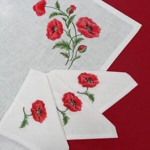 May include: White linen napkins with red poppy embroidery. The napkins are folded in a triangle shape and are arranged on a red surface. The embroidery is detailed and realistic, with green leaves and stems.