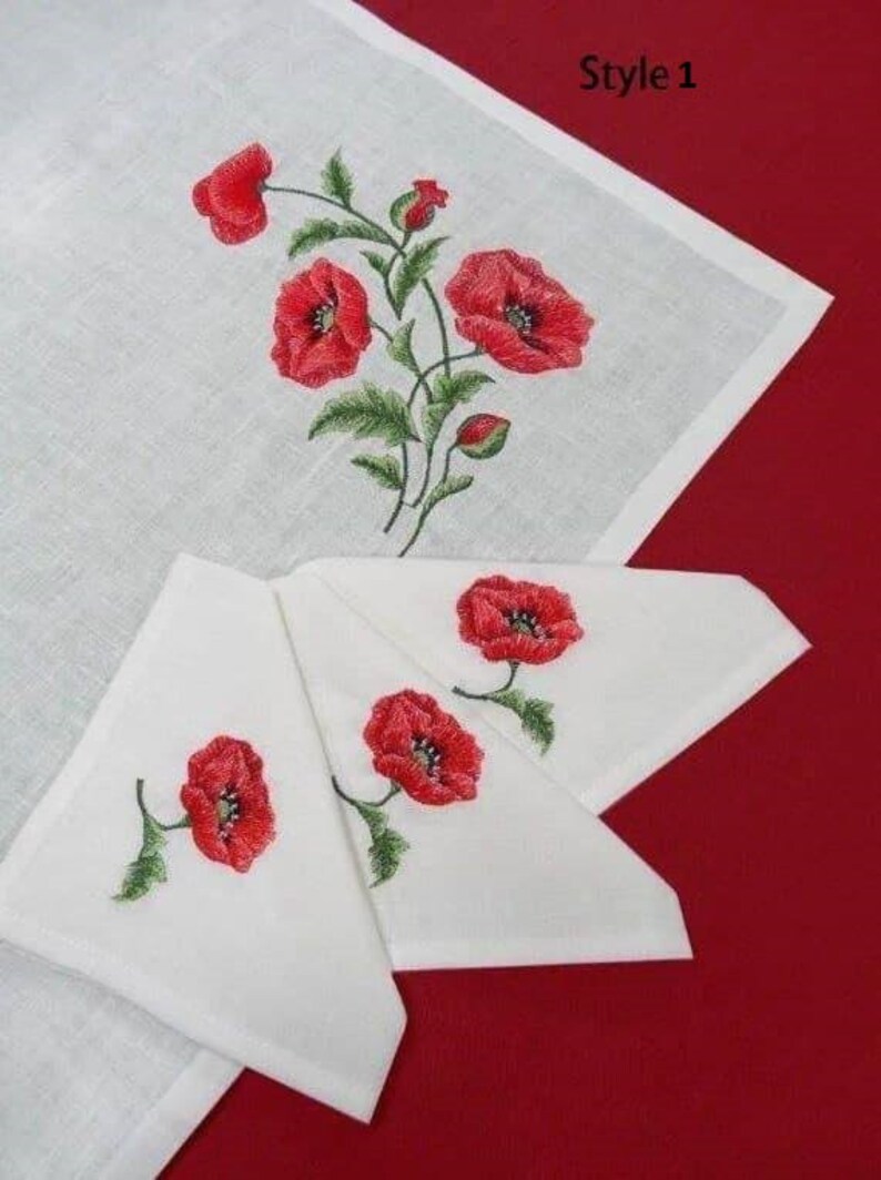 May include: White linen napkins with red poppy embroidery. The napkins are folded in a triangle shape and are arranged on a red surface. The embroidery is detailed and realistic, with green leaves and stems.