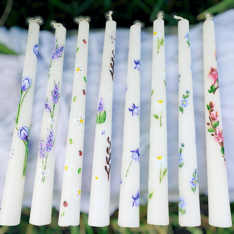 May include: A collection of white taper candles, each hand-painted with floral designs. The candles feature various flowers in shades of purple, blue, pink, and yellow, with green leaves and accents. The candles are arranged closely together, creating a visually appealing display.