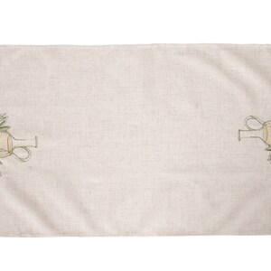 May include: A beige linen table runner with a green and yellow embroidered design of a pitcher and olive branches on each end.