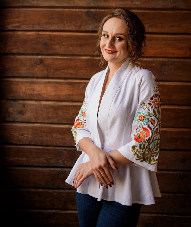 Op de afbeelding: Een vrouw draagt een witte linnen blouse met geborduurde bloemenmotieven op de mouwen. De blouse heeft een losse pasvorm en een V-hals. Ze staat voor een houten muur.