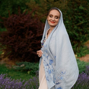 Puede incluir: Un chal de lino azul claro con dise&ntilde;os florales azules bordados. El chal est&aacute; drapeado sobre un vestido beige. El fondo presenta follaje verde y un campo de lavanda, creando una est&eacute;tica serena y natural.