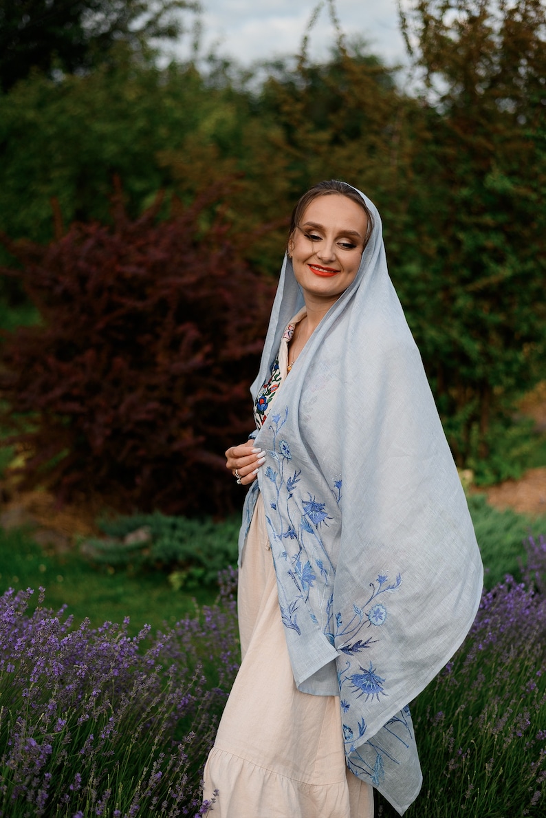 Puede incluir: Un chal de lino azul claro con dise&ntilde;os florales azules bordados. El chal est&aacute; drapeado sobre un vestido beige. El fondo presenta follaje verde y un campo de lavanda, creando una est&eacute;tica serena y natural.