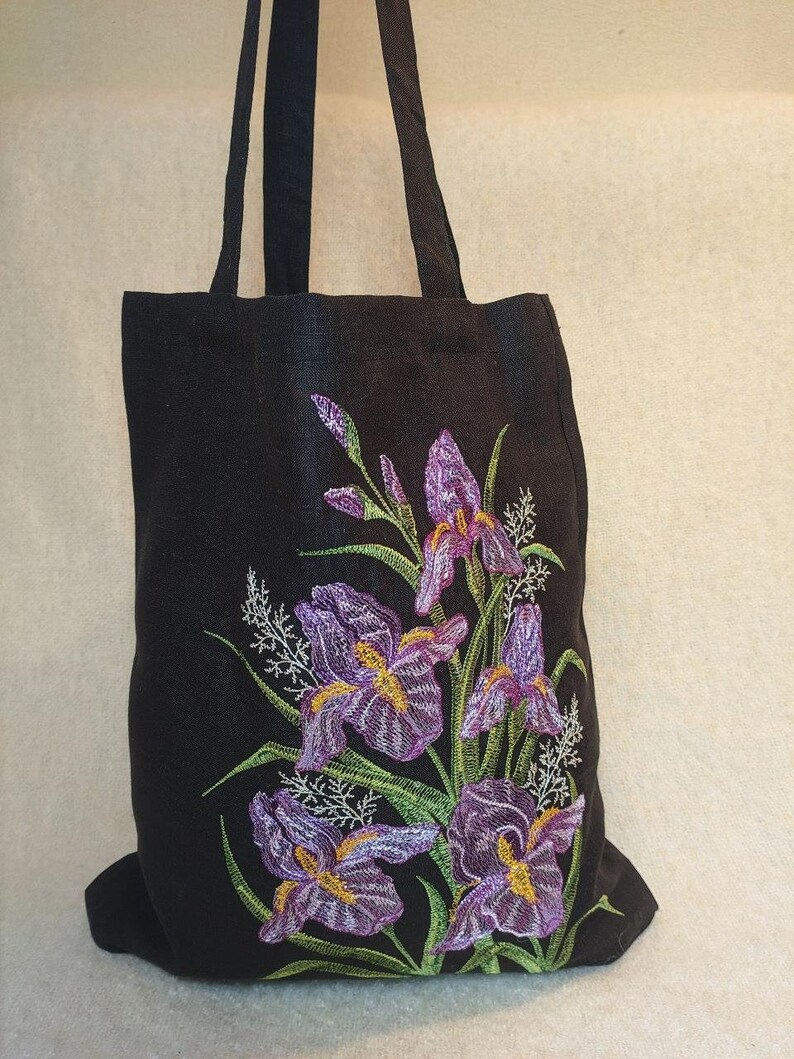 May include: Black tote bag featuring an embroidered design of purple iris flowers with green leaves and stems.