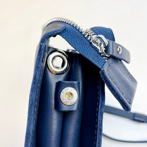 May include: A navy blue leather crossbody bag with a zipper closure and a silver metal ring. The bag has a flap with a silver metal button.
